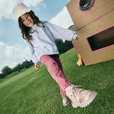 Niña con botas oro rosado Coqueta y Audaz caminando sobre césped junto a un cohete de cartón. Colección Pisa Fuerte, libertad y estilo en cada paso.